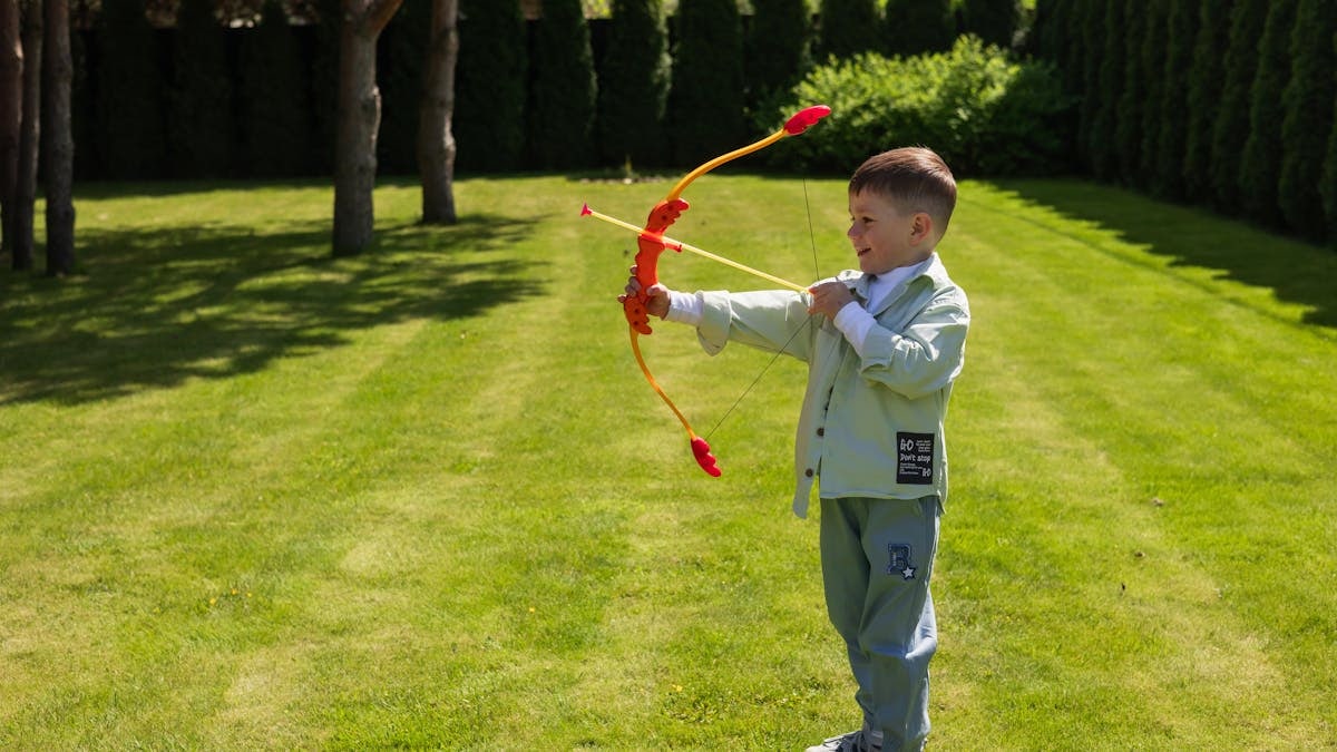Young boy enjoying a sunny day outside, aiming with a colorful toy bow in a lush garden.